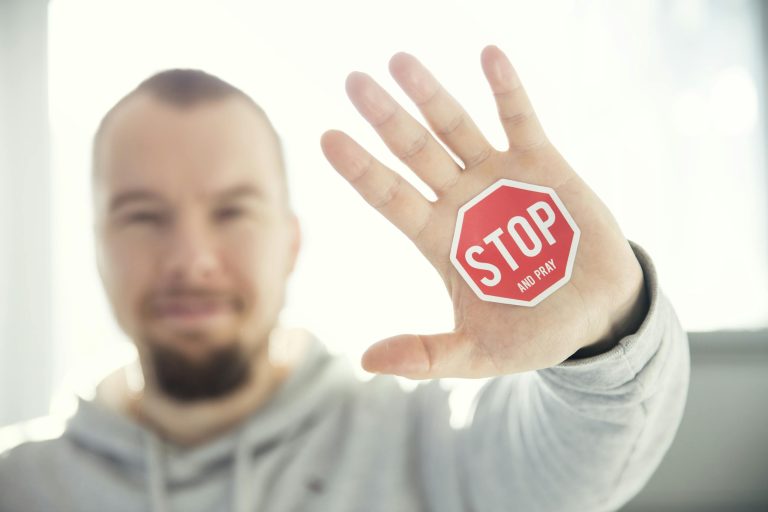 A man in a hoodie holds up his hand with a 'Stop and Pray' sticker. Focus on hand and sticker.