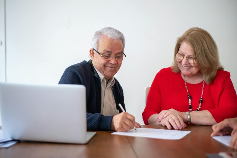 Elderly couple signing documents at a desk with a laptop. Smiling and working.